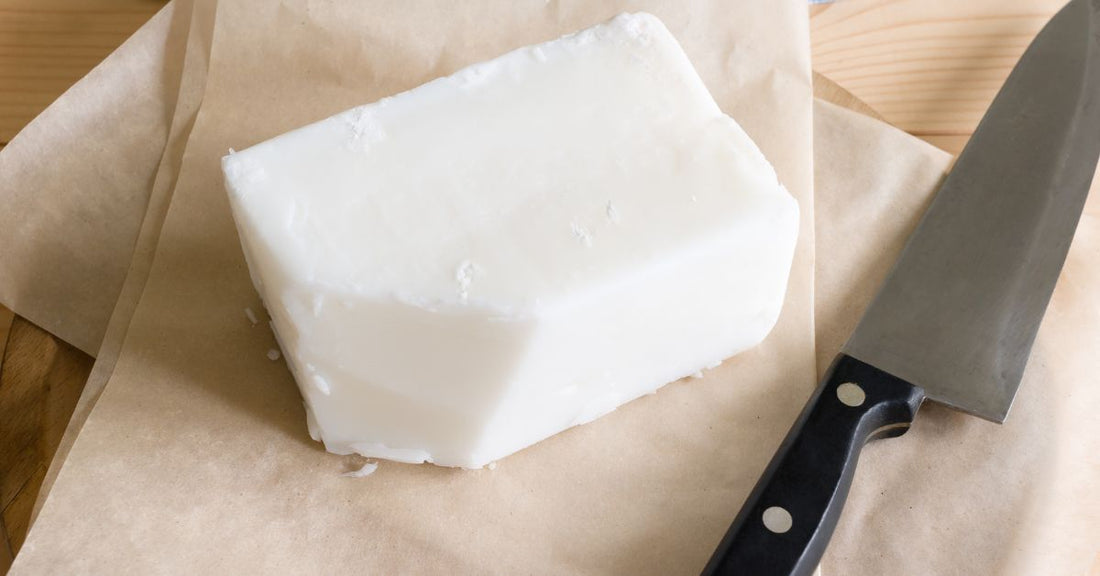 A block of white beef tallow sitting on parchment paper beside a knife. A corner of the block has been cut off.