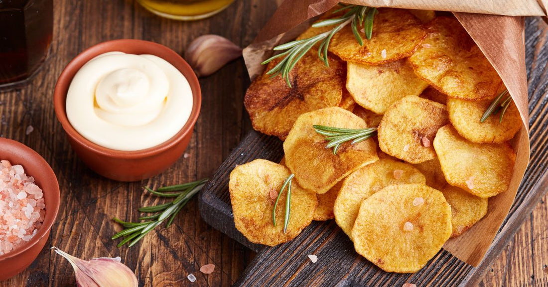 Crisp potato chips wrapped in paper and displayed on a wooden charcuterie board. There are bowls of salt and dip beside it.