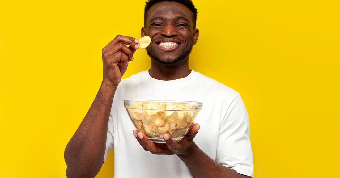A man smiling as he holds a bowl of potato chips in one hand and a single chip up to his face with the other.